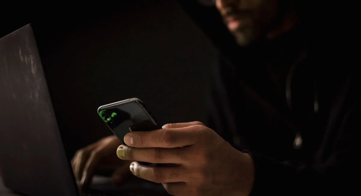 Crop anonymous ethnic male cyber spy with cellphone and netbook hacking system in evening