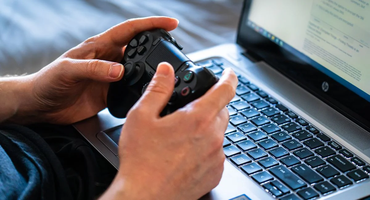 Person holds a gaming controller while interacting with a laptop, emphasizing modern gaming technology.