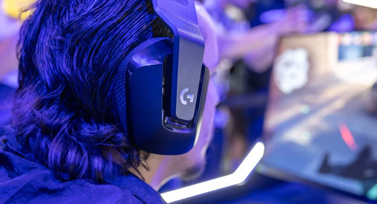 Focused gamer with headphones playing a video game on a PC under blue lighting in a crowded room.