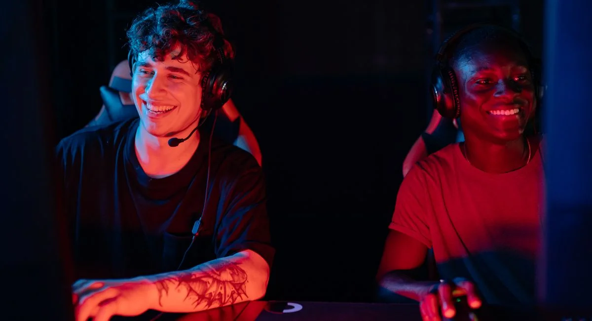 Smiling male gamers playing on PC with headsets in a dimly lit room.