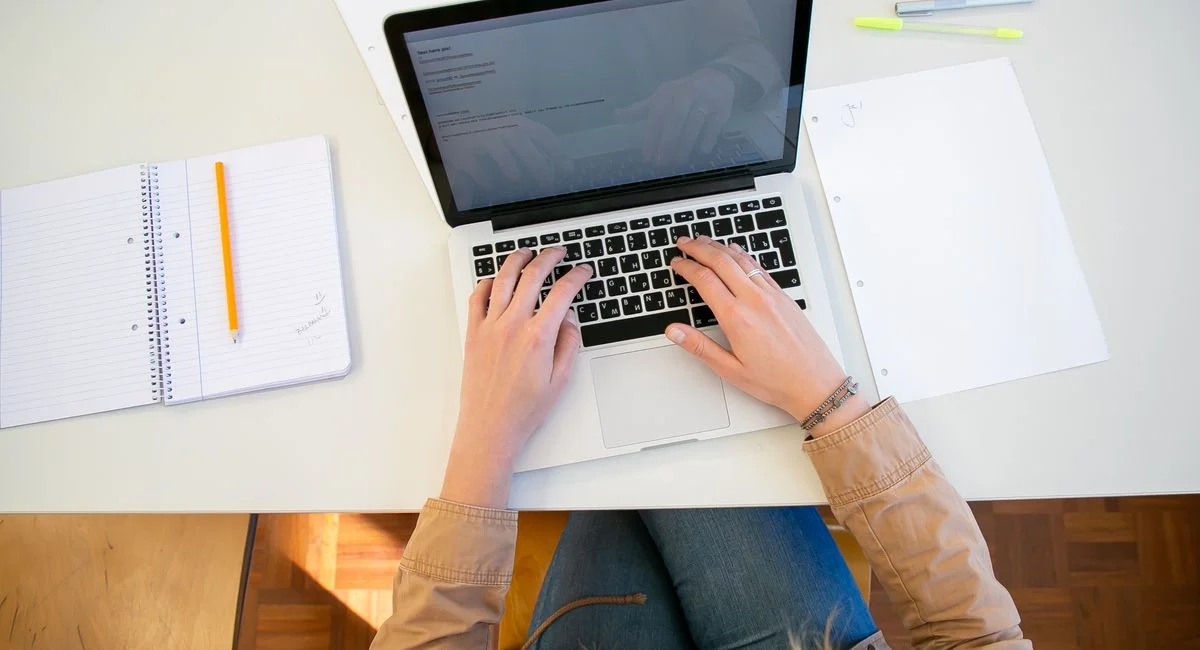 Top view of crop anonymous female student typing on keyboard while preparing for university assignment at table with notebook